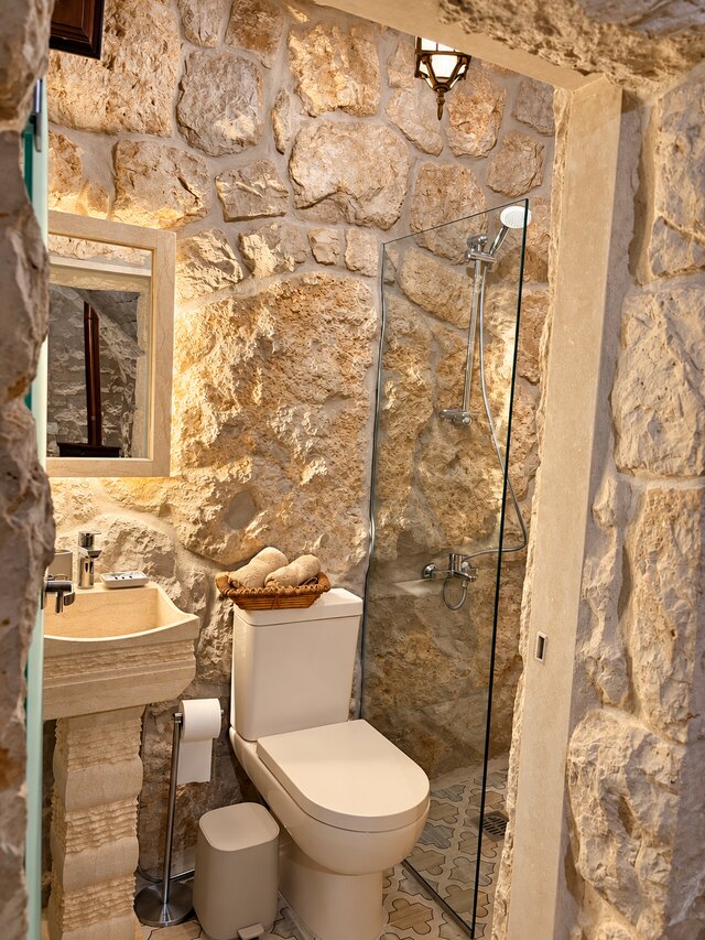 Gemelli's ensuite bathroom, glass shower panel against the stone wall beside the sink and toilet.